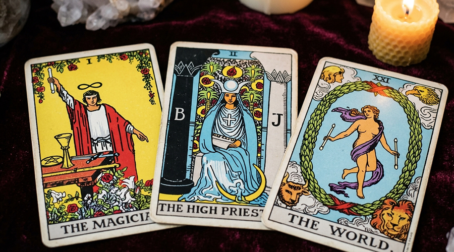 A close-up of The Magician, The High Priestess, and The World Tarot cards resting on purple velvet next to a lit candle